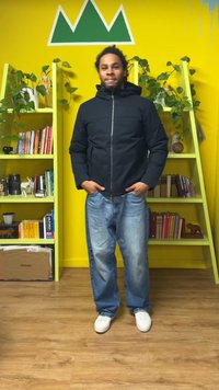 Μαύρο μπουφάν με επένδυση και φερμουάρ μπροστά, φορεμένο πάνω από ανοιχτόχρωμα μπλε τζιν και λευκά αθλητικά παπούτσια. Το φόντο περιλαμβάνει έναν κίτρινο τοίχο και πράσινα ράφια.