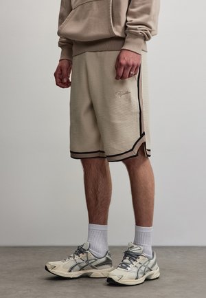 Mand iført beige teksturerede shorts med sidelængder, hvide crew-sokker og beige atletiksneakers står på en grå gulv.