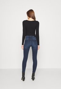 Mujer de espaldas vistiendo una camiseta de manga larga negra, jeans ajustados de color azul oscuro y botas negras de tobillo, con fondo blanco liso.