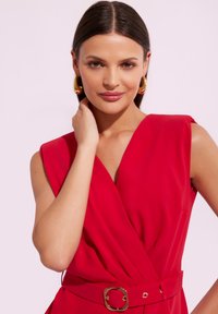 Czerwona sukienka bez rękawów z głębokim dekoltem w kształcie litery V i dopasowanym stanem, z złotym zapięciem na pasie w tym samym kolorze. Gładka faktura materiału.