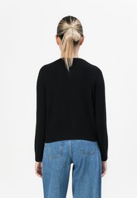 Mulher com cabelo loiro preso em um coque, vestindo uma blusa de manga longa preta e jeans azuis, de pé voltada para longe contra um fundo branco.