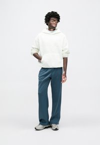 Sweat à capuche vert clair avec un motif texturé, associé à un pantalon bleu sarcelle foncé et des baskets grises. La tenue présente une coupe décontractée.