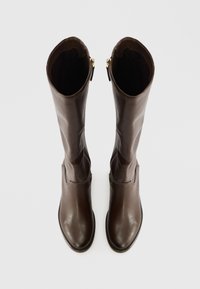 Brązowe skórzane kozaki do kolan o gładkiej fakturze, zaokrąglonym nosku i falistym górnym brzegu. Posiadają boczny zamek błyskawiczny ze złotym wykończeniem.