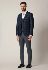 Granatowa marynarka w kratę o dopasowanym kroju, zestawiona z szarym kamizelką i spodniami. Biała koszula z kołnierzykiem, uzupełniona czarnymi butami.