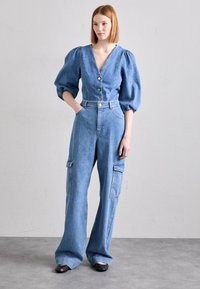 Denim jumpsuit med puffärmar, djup V-ringning, tre knappar fram, vida ben och sidofickor i cargo-stil i ljusblått.