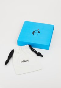Caja azul brillante con texto negro que presenta la letra "é" y un acento rojo. Bolsa blanca con cordones negros y "élio" impreso en negro.
