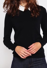 Svart stickad tröja med ribbad halsringning och ärmar, med en vit spetskrage under. Slät textur, figursydd design.