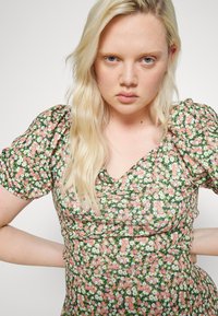 Vestido floral com fundo verde e flores rosas e brancas. Mangas bufantes e design em V. Tecido suave com textura plissada.