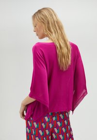 Poncho en tricot fuchsia avec des manches larges, texture légèrement transparente et encolure arrondie, associé à une jupe multicolore à motifs.