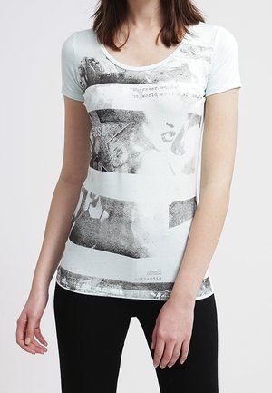 Kobieta ubrana w jasnoniebieską dopasowaną koszulkę z wyblakłym czarno-białym abstrakcyjnym nadrukiem oraz czarne spodnie, stojąca na jednolitym tle.