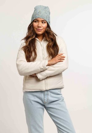 Giacca puffa leggera con zip frontale, tasche laterali e berretto a coste blu. Il modello posa con le braccia incrociate, indossando jeans chiari.