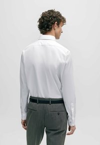 Chemise blanche à manches longues avec un col classique, associée à un pantalon rayé gris et une ceinture noire, vue de dos.