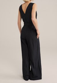 Czarny kombinezon o bezrękawnikowym kroju, z dekoltem w kształcie litery V z tyłu, paskiem w talii oraz szerokimi nogawkami. Wykonany z fakturowanej tkaniny o matowym wykończeniu.