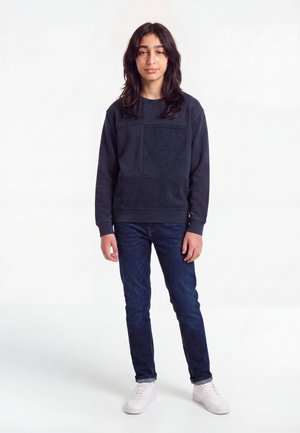 Adolescent aux longs cheveux foncés portant un sweat-shirt noir Guess, un jean bleu foncé et des baskets blanches sur un fond blanc uni.