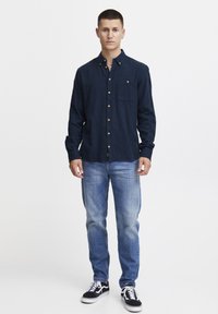 Temno modra srajca s kratkimi rokavi in žepom, v kombinaciji s svetlomodrimi jeans hlačami in črnimi supergami. Ima zlato gumbe in dolge rokave.