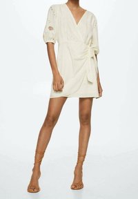 Vestido cruzado crema con mangas abullonadas y bordado de ojales. Cuenta con un lazo en la cintura y un dobladillo corto. Combinado con sandalias de tiras en color camel.