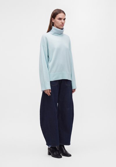 Femme portant un pull à col roulé bleu clair, un pantalon large bleu foncé et des bottines noires, debout devant un fond blanc uni.