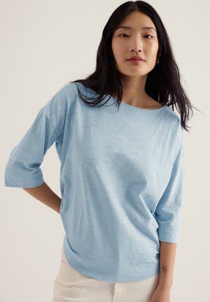 Kobieta o długich ciemnych włosach, ubrana w jasnoniebieską bluzkę z rękawem do łokcia i kremowe spodnie, stojąca z jedną ręką na biodrze.