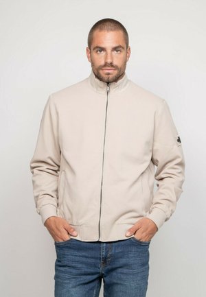 Homme aux cheveux courts et à la barbe porte une veste zippée beige clair et un jean bleu, debout les mains dans les poches devant un fond uni.