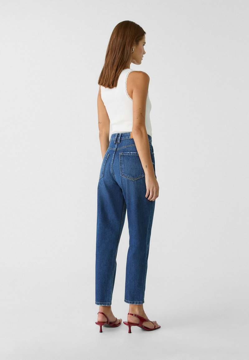 Hochtaillierte blaue Denim-Jeans mit geradem Bein, ausgestattet mit zwei Gesäßtaschen und einem ausgefransten Saum, kombiniert mit roten Sandalen mit Absatz.