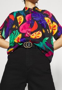 Camicia nera a maniche corte con vivaci motivi di frutta e tropicali in rosa, giallo, verde e viola, con bottoni marroni e colletto.