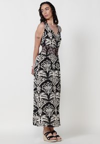 Robe maxi florale noire et blanche avec un décolleté en V et une taille smockée. Tissu lisse avec des motifs de feuilles détaillés, portée avec des sandales compensées noires.