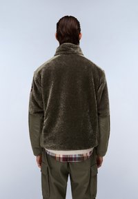 Oliviavihreä fleece-takki, jossa on korkea kaulus, teksturoitua kangasta ja korostetut hihat; päällä ruudullinen paita ja cargo-housut.