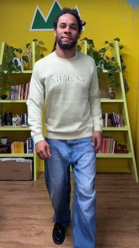 Lysegrøn sweatshirt med præget logo, kombineret med løstsiddende blå jeans og sorte sko; står i et rum med gule vægge og hylder.