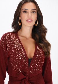 Hosszú barna hullámos hajú nő arany fülbevalóval és mélyvörös flitteres kardigánnal, amelyet a derékban kötött, fekete felsőre.