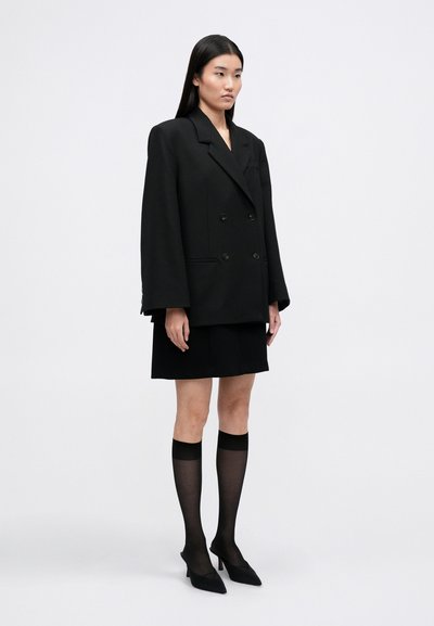 Blazer preto de duas fileiras de botões com lapelas recortadas, uma silhueta fluida, combinado com uma saia preta e meias altas transparentes até ao joelho. Sapatos de salto alto pretos com bico.