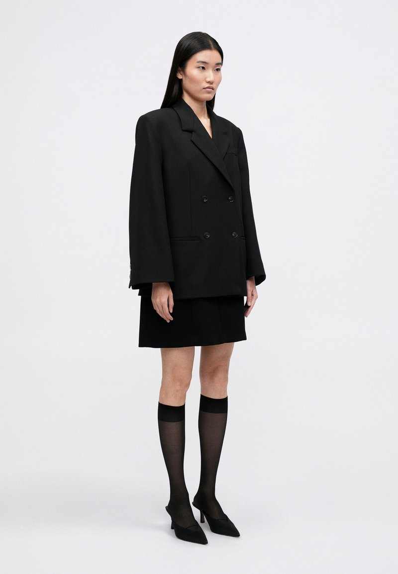 Black double-breasted blazer with notched lapels, a flowing silhouette, paired with a black skirt and sheer knee-high socks. Pointed black heels.