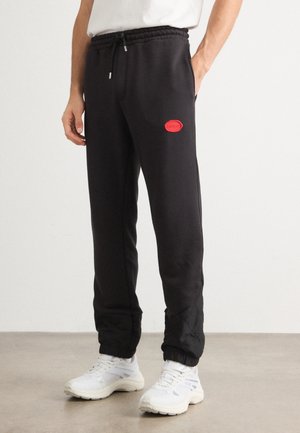 Mann trägt schwarze Jogger-Hose mit rotem Logo am Oberschenkel und weißen Sneakern, steht vor schlichtem hellem Hintergrund.