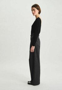 Haut noir à manches longues associé à un pantalon gris à rayures fines taille haute, avec une coupe décontractée et une texture lisse. Des chaussures noires complètent le look.