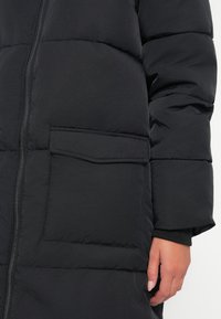 Schwarze Pufferjacke mit Reißverschluss vorne, ausgestattet mit einer quadratischen Tasche auf der linken Seite. Der Stoff hat eine glatte Textur mit genähten Abschnitten.