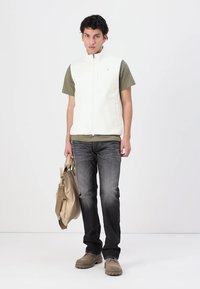 Weißer Zip-Up-Weste über einem olivgrünen T-Shirt, kombiniert mit dunkelgrauen Jeans und beigen Schuhen. Hält eine beige Tasche und steht vor einem schlichten Hintergrund.