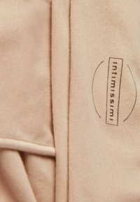 Lichtbeige stof met een zachte textuur, voorzien van een prominent gedrukt logo met de tekst "intimissimi." Eenvoudig ontwerp met subtiele stikseldetails.