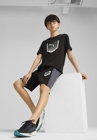 T-shirt nera a maniche corte con un logo grafico argento, abbinata a pantaloni corti neri con un pannello grigio e logo. Sneakers nere con accenti turchesi.