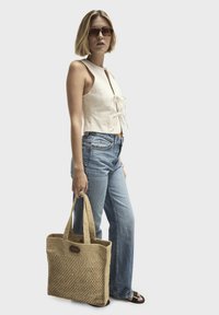 Borsa a rete leggera beige chiaro con motivo testurizzato, manici robusti e un accento in pelle marrone. Modello abbinato a jeans casual e una canotta.