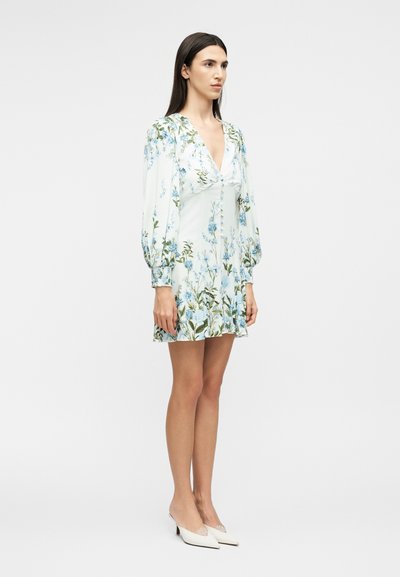 Femeie în picioare purtând o rochie mini albă cu mâneci lungi și model floral, cu nasturi în față și pantofi cu toc ascuțit albi, pe un fundal simplu.