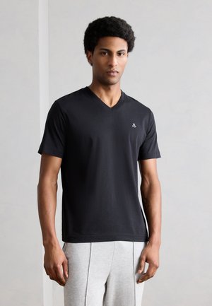 Młody mężczyzna z krótkimi kręconymi włosami, ubrany w czarną koszulkę z dekoltem w serek z małym białym logo oraz jasnoszare spodnie, stoi na tle szarej ściany.