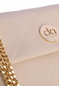 Bolso de mano beige de piel sintética acolchada con una correa de cadena dorada y un emblema de logotipo circular en el frente. Costuras detalladas visibles.