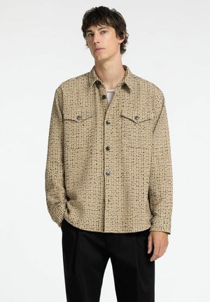 Beige skjorte med et struktureret mønster, to brystlommer og knaplukning. Lange ærmer, afslappet pasform, parret med sorte bukser.