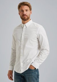 Camisa blanca de botones hecha de algodón con una textura sutil. Presenta un cuello clásico, botones amarillos y un logo bordado en el pecho.