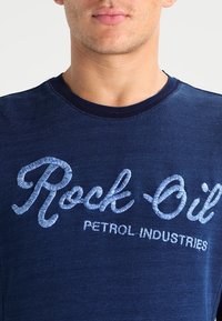 Námořnická modrá tričko s kulatým výstřihem, obsahující světle modrý vyšívaný text "Rock Oil" a "PETROL INDUSTRIES," texturovaná tkanina a moderní střih.
