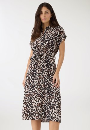 Donna che indossa un vestito leopardato a maniche corte fino al ginocchio, con bottoni e cintura annodata in vita, in piedi davanti a uno sfondo semplice.