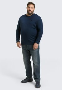 Dunkelblauer Strickpullover mit langen Ärmeln und Rundhalsausschnitt, kombiniert mit ausgebleichten blauen Jeans und schwarzen Turnschuhen. Einfaches Design, keine sichtbaren Muster.