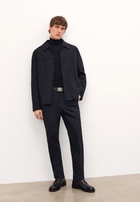 Chaqueta de mezcla de lana negra con corte recto, cierre de botones y un acabado texturizado, combinada con pantalones a medida a juego y zapatos negros.