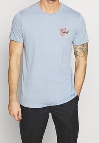 Světle modré bavlněné tričko s kulatým výstřihem, krátkými rukávy a malou růžovou grafiku donutu s textem "Rychlé sousto" na levém prsu.