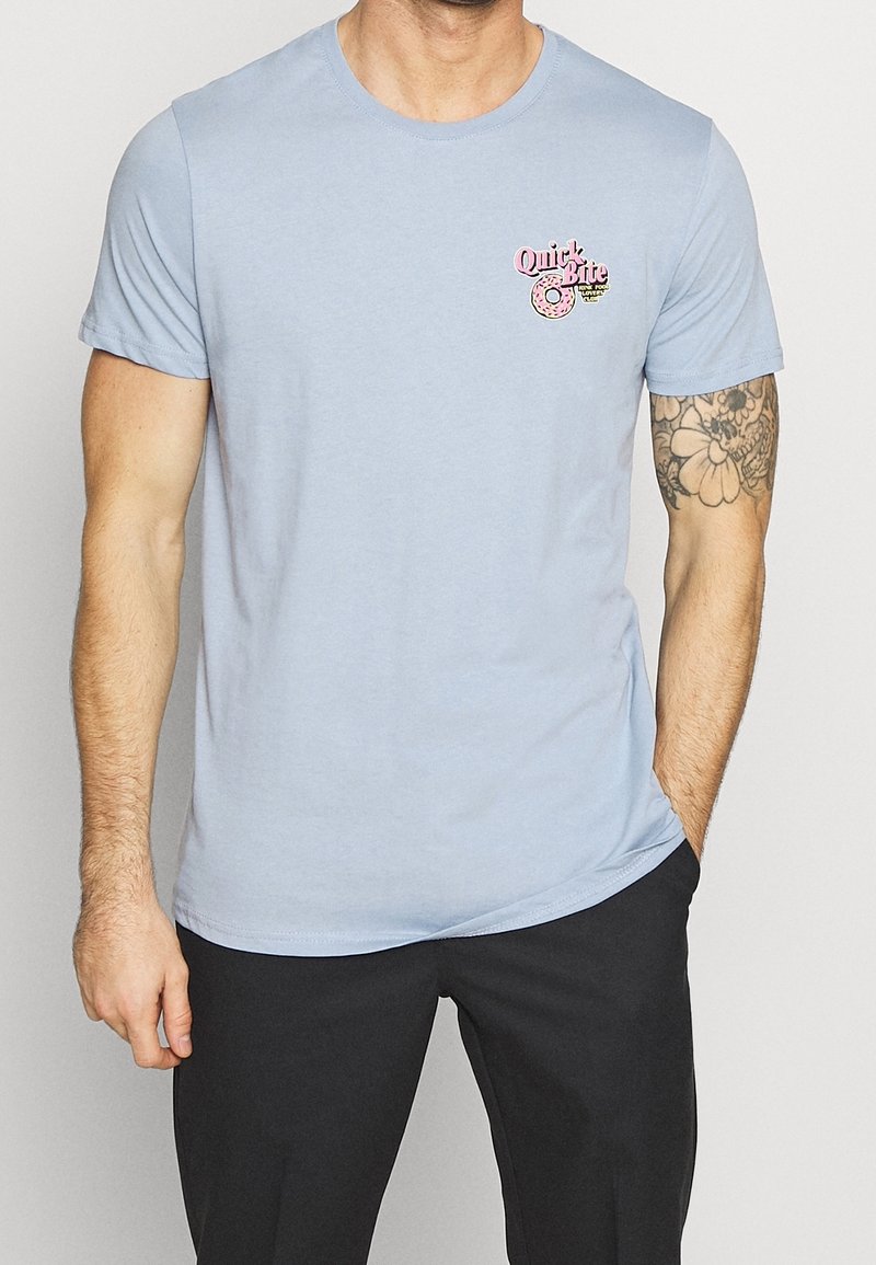 Světle modré bavlněné tričko s kulatým výstřihem, krátkými rukávy a malou růžovou grafiku donutu s textem "Rychlé sousto" na levém prsu.