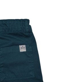 Teal-färbede Stoffshorts mit einer Gesäßtasche und einem hellgrauen Etikett, das ein gekreuztes Gabel- und Messerdesign zeigt. Glatte Textur, strapazierfähiges Material.
